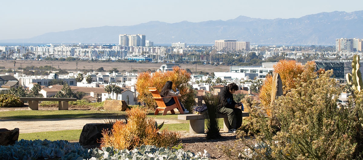 Two students sitting on benches near the bluff with mountains, Playa Vista, and the ocean in the distance