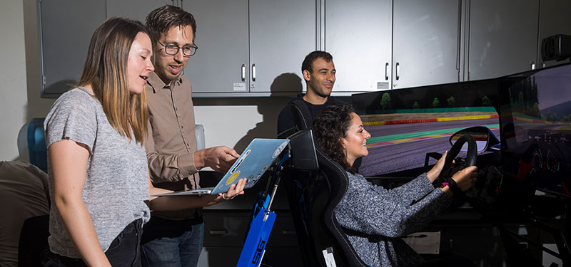 A student driving in a car physics simulator with several other students watching