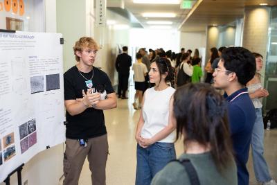 Group shot of students. Male student standing in front of poster explaining his work to other students.